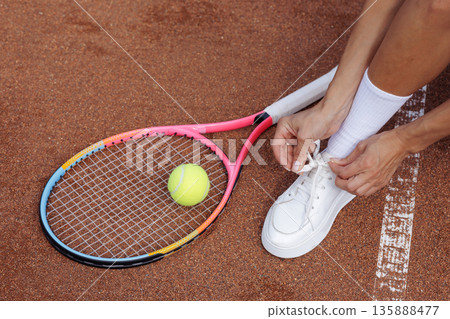 Closeup of female tennis player tying shoelace beside racket and ball on clay court, ideal for sportswear website banner or training blog. Closeup of female tennis player tying shoelace beside racket and ball on clay court, ideal for sportswear website banner or training blog. 135888477