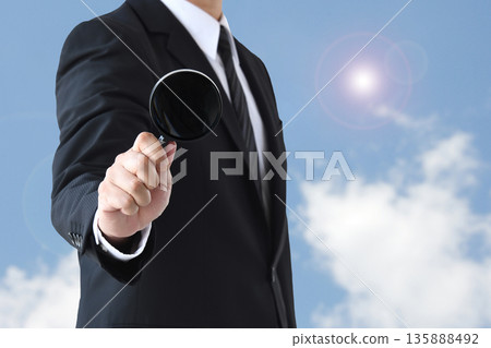 Businessman holding a magnifying glass against the blue sky 135888492