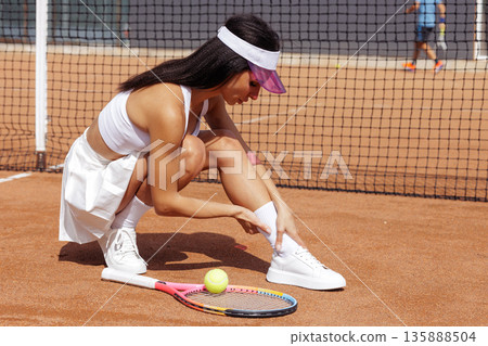 Female tennis athlete crouching with ankle pain on outdoor clay court, racket and ball nearby. For sports medicine, injury prevention, and training content on websites or ads. Female tennis athlete crouching with ankle pain on outdoor clay court, racket and ball nearby. For sports medicine, injury prevention, and training content on websites or ads. 135888504