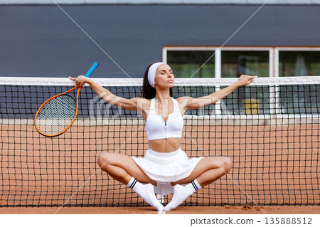 Athletic woman in white tennis outfit stretches at the net on a clay court. Suitable for website banner or social ad about tennis training, fitness, or wellness. 135888512
