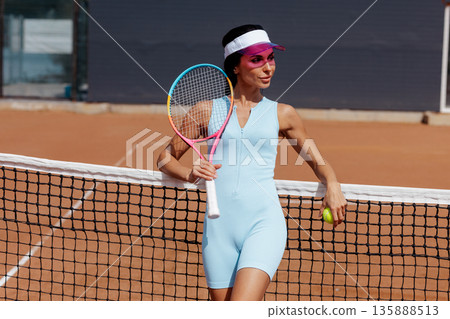 Athletic woman holding tennis racket and ball by the net on an outdoor clay court, ideal for website banner or sports advertising. 135888513