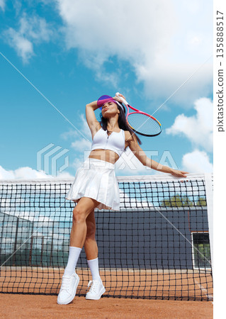 Full body female tennis player posing by the net on an outdoor clay court, with copy space sky for website banner or social ad. Full body female tennis player posing by the net on an outdoor clay court, with copy space sky for website banner or social ad. 135888517