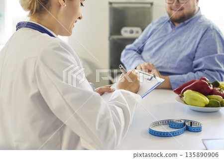 Nutritionist consulting overweight patient with vegetables, tape measure in clinic Nutritionist consulting overweight patient with vegetables, tape measure in clinic 135890906