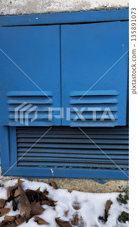 Close-up of a blue vent cover surrounded by snow and dry leaves. Close-up of a blue vent cover surrounded by snow and dry leaves. 135891073