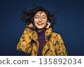 AI-generated photo of a smiling young Japanese woman wearing a yellow down jacket against a navy blue background 135892034