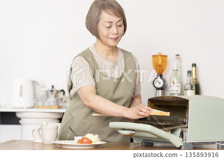 Senior woman toasting bread and eating breakfast in the kitchen Senior woman toasting bread and eating breakfast in the kitchen 135893916