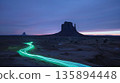 A lone car winds its way through the desert leaving behind a glowing green trail of light near a rugged monolith 135894448