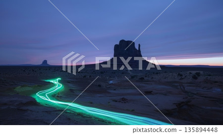 A lone car winds its way through the desert leaving behind a glowing green trail of light near a rugged monolith 135894448
