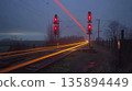 The humble train crossing in the rural countryside becomes a picturesque moment with light trails from passing cars 135894449