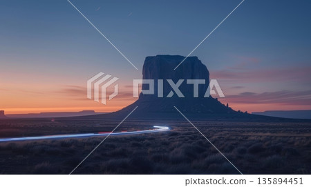 As the sun sets behind a massive desert monolith cars can be seen speeding by leaving behind a trail of shimmering blue light 135894451