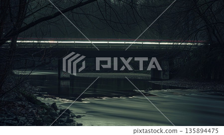 A peaceful sight of a bridge over a tranquil river with the faint lights of cars creating subtle light trails in the darkness adding a touch of mystery to the scene 135894475