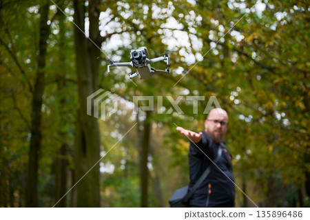In a lush forest during autumn, a man extends his hand as a drone takes flight, exploring the rich colors of trees around him. Technology meets nature in harmony. 135896486
