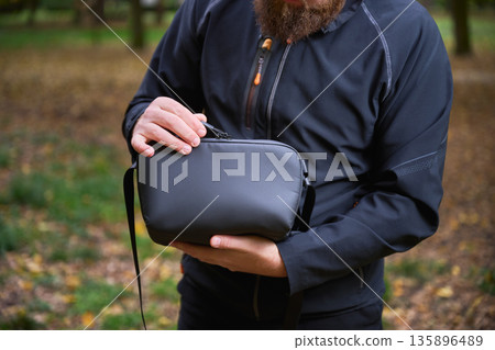 A man stands in a peaceful park, cradling a stylish bag close to his chest. Autumn leaves surround him as he enjoys the serene atmosphere of the afternoon. A man stands in a peaceful park, cradling a stylish bag close to his chest. Autumn leaves surround him as he enjoys the serene atmosphere of the afternoon. 135896489