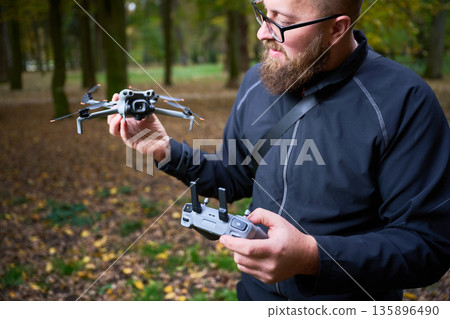 A bearded man in a black jacket holds a drone and controller in a colorful forest. Bright leaves surround him as he prepares for an exciting flight. A bearded man in a black jacket holds a drone and controller in a colorful forest. Bright leaves surround him as he prepares for an exciting flight. 135896490