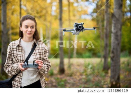 In a vibrant autumn forest, a young woman controls a drone with enthusiasm while surrounded by golden leaves and tall trees. 135896519