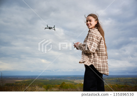 A young girl stands on a hilltop, enjoying the fresh air while skillfully operating a drone. The windy weather adds excitement to her adventure. A young girl stands on a hilltop, enjoying the fresh air while skillfully operating a drone. The windy weather adds excitement to her adventure. 135896530