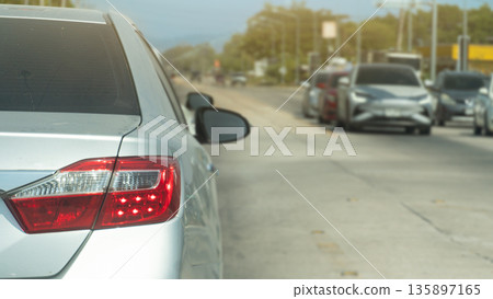 Rear side of silver car with turn on brake light on concrete road. Cars are parked waiting at the traffic light on the opposite side, despite the clear traffic conditions. 135897165