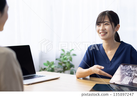 A young woman wearing medical scrubs talking to a patient while holding a tablet displaying an X-ray image 135897247