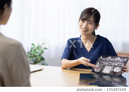 A young woman wearing medical scrubs talking to a patient while holding a tablet displaying an X-ray image 135897249