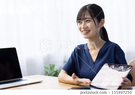 一位身穿醫用工作服的年輕女子一邊與病人交談,一邊手持一台顯示X光影像的平板電腦。 一位身穿醫用工作服的年輕女子一邊與病人交談,一邊手持一台顯示X光影像的平板電腦。 135897250