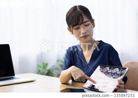 一位身穿醫用工作服的年輕女子一邊與病人交談，一邊手持一台顯示X光影像的平板電腦。 135897251