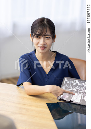 一位身穿醫用工作服的年輕女子一邊與病人交談，一邊手持一台顯示X光影像的平板電腦。 135897252