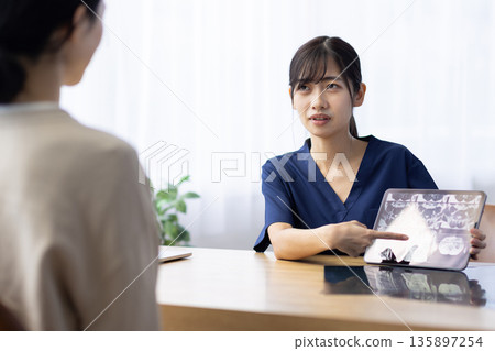 A young woman wearing medical scrubs talking to a patient while holding a tablet displaying an X-ray image A young woman wearing medical scrubs talking to a patient while holding a tablet displaying an X-ray image 135897254