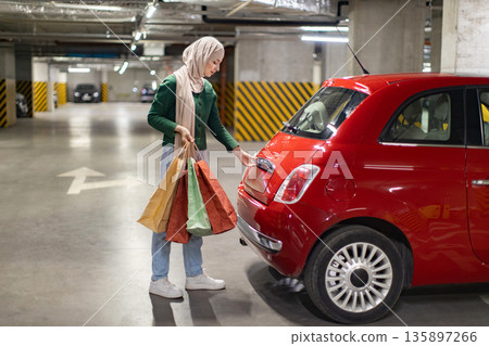 A woman wearing a hijab prepares to load shopping bags into the trunk of her red car in a parking garage A woman wearing a hijab prepares to load shopping bags into the trunk of her red car in a parking garage 135897266