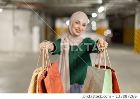 A smiling woman in a hijab holds up shopping bags in a parking garage, ready to show off her purchases A smiling woman in a hijab holds up shopping bags in a parking garage, ready to show off her purchases 135897269
