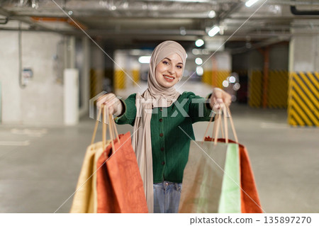 A smiling woman in a hijab holds up shopping bags in a parking garage A smiling woman in a hijab holds up shopping bags in a parking garage 135897270