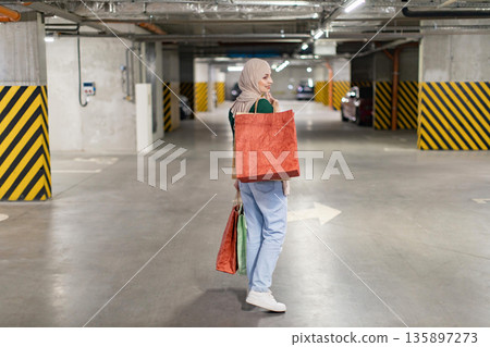 A woman wearing a hijab carries shopping bags in an underground parking garage, looking back over her shoulder A woman wearing a hijab carries shopping bags in an underground parking garage, looking back over her shoulder 135897273
