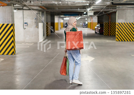 A woman wearing a hijab carries shopping bags in an underground parking garage, likely after a successful shopping trip A woman wearing a hijab carries shopping bags in an underground parking garage, likely after a successful shopping trip 135897281