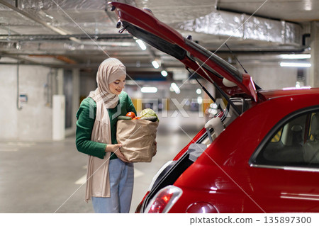 A woman wearing a hijab smiles while putting groceries into the trunk of her red car in a parking garage A woman wearing a hijab smiles while putting groceries into the trunk of her red car in a parking garage 135897300