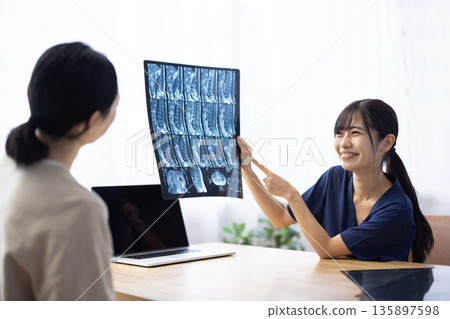 A young Japanese woman wearing a navy blue medical scrub explaining to a patient while showing him an X-ray 135897598