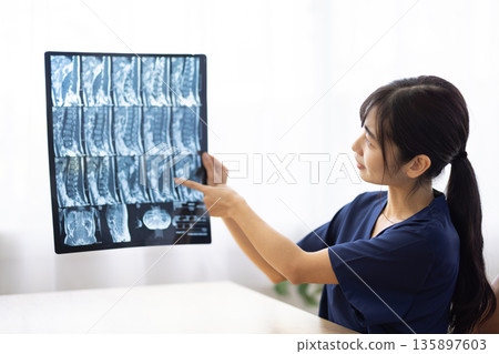 一位身穿藏藍色醫用工作服的年輕日本女性正在向病人展示X光片並進行講解。 135897603