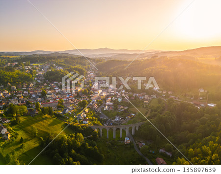 The railway bridge arches over the town of Smrzovka as the sun sets in the background. Houses are scattered throughout the green landscape, highlighting the rural setting. The railway bridge arches over the town of Smrzovka as the sun sets in the background. Houses are scattered throughout the green landscape, highlighting the rural setting. 135897639