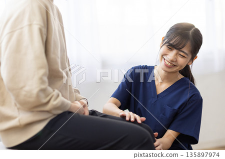 一位身穿藏藍色手術服的年輕女子麵帶微笑,一邊把手放在病人膝蓋上一邊與他交談。 一位身穿藏藍色手術服的年輕女子麵帶微笑,一邊把手放在病人膝蓋上一邊與他交談。 135897974