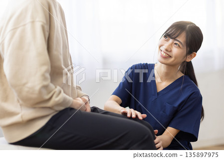一位身穿藏藍色手術服的年輕女子麵帶微笑,一邊把手放在病人膝蓋上一邊與他交談。 一位身穿藏藍色手術服的年輕女子麵帶微笑,一邊把手放在病人膝蓋上一邊與他交談。 135897975