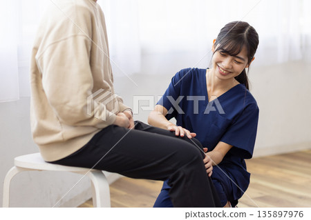 A young woman wearing a navy blue scrub smiles and talks with her hand on the patient's knee A young woman wearing a navy blue scrub smiles and talks with her hand on the patient's knee 135897976
