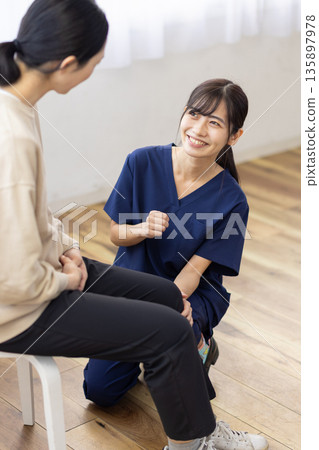 A young woman wearing a navy blue scrub smiles and talks with her hand on the patient's knee A young woman wearing a navy blue scrub smiles and talks with her hand on the patient's knee 135897978