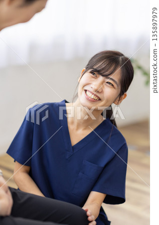 A young woman wearing a navy blue scrub smiles and talks with her hand on the patient's knee A young woman wearing a navy blue scrub smiles and talks with her hand on the patient's knee 135897979