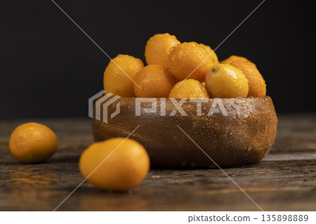 wet small orange fruits of juicy fresh and fragrant kumquat on a wooden table, oval oblong kumquat fruit covered with drops of water and scattered on a black table made of pine wood wet small orange fruits of juicy fresh and fragrant kumquat on a wooden table, oval oblong kumquat fruit covered with drops of water and scattered on a black table made of pine wood 135898889