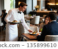 Professional Male Waiter in White Uniform and Bowtie Serving Plates of Gourmet Food to Guests in a Luxury Restaurant. High-End Fine Dining Hospitality and Service Concept. 135899610