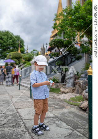 Young Boy Using Toy Camera to Photograph Statues in Lush Garden Setting in Bangkok, Thailand 135900900
