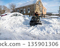Man on ATV plows snow clearing driveway with a car buried in snowdrift 135901306