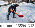 A man in a snowsuit and fur hat shovels snow on a rural farm landscape with street 135901312