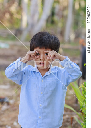 Portrait of happy cute Asian Thai little boy rounding his eyes with his fingers in the garden 135902604
