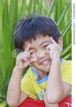 Portrait of happy cute Asian Thai little boy rounding his eyes with his fingers sitting in the park 135902864