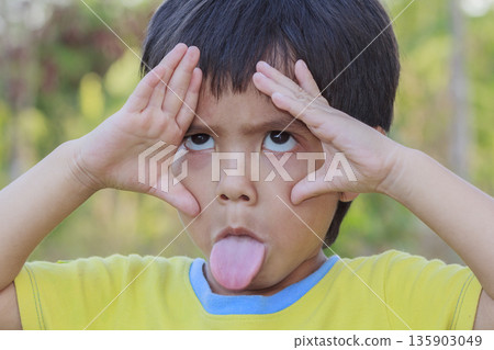Portrait of happy cute Asian Thai little boy rounding his eyes with his fingers and sticking his tongue out outdoor Portrait of happy cute Asian Thai little boy rounding his eyes with his fingers and sticking his tongue out outdoor 135903049