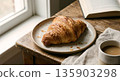 Crispy croissant on a plate next to a cup of coffee and an open book on a rustic wooden table by the window 135903298